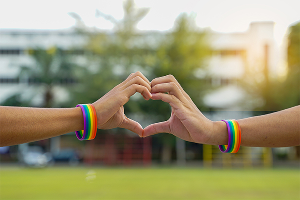 Two people, each wearing a rainbow pride wristband putting their hands together to make a heart shape.