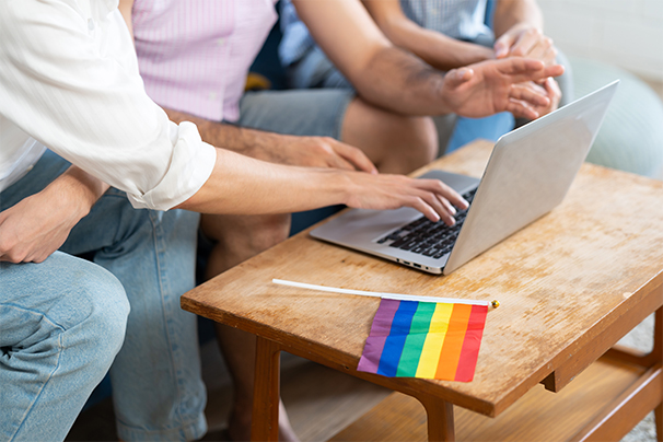 People gathered round a laptop which is sitting on a table next to a pride flag.
