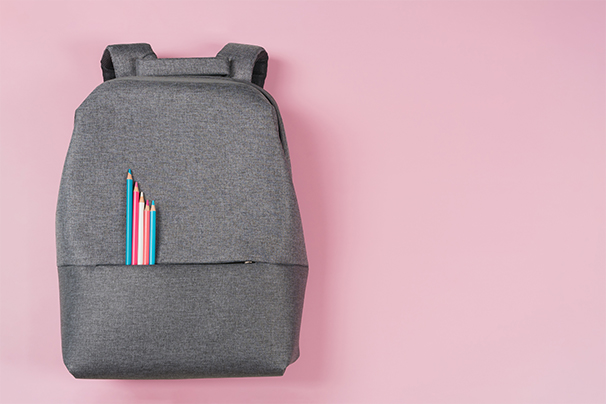 A grey backpack on a pink background with a collection of pencils showing in the front pocket.