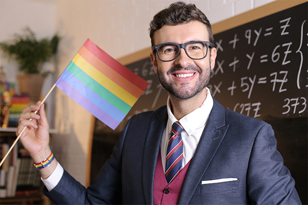 An individual wearing a blue suit and glasses, holding a pride flag and smiling.