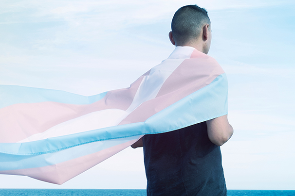 A person looking out towards the ocean wrapped in a transgender pride flag.