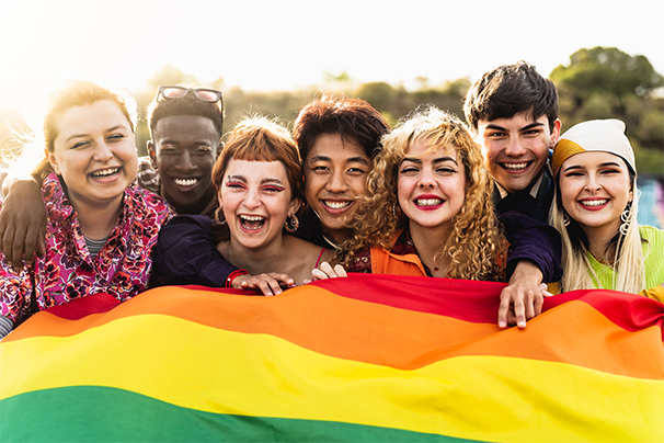 A group of happy LGBTQ+ youth standing behind a large pride flag.